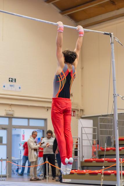 Young male gymnast hangs from the high bar, arms extended overhead, wearing a red and grey competition leotard.