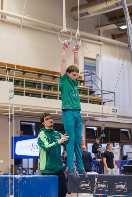 Young gymnast hangs from still rings while his coach watches attentively with hands clasped at a Gymfed competition.