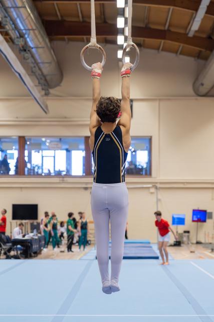 Male gymnast hangs from still rings, arms extended overhead, in a bright indoor gymnastics hall with teammates visible behind.