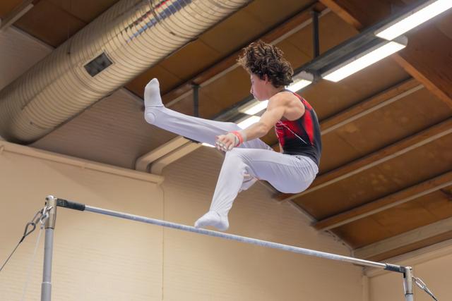 Young male gymnast grips the high bar mid-routine, legs tucked upward with intense focus in an indoor gymnasium.
