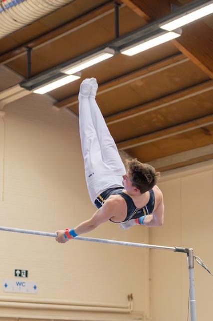 Young male gymnast performs an inverted handstand on the horizontal bar, body fully extended, in an indoor gymnasium.
