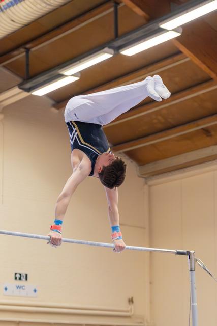 Young male gymnast performing an inverted handstand on the horizontal bar, body fully extended upward in an indoor gym.