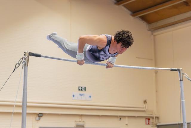 Young male gymnast grips the high bar with fierce concentration, body extended horizontally in a dynamic hold.