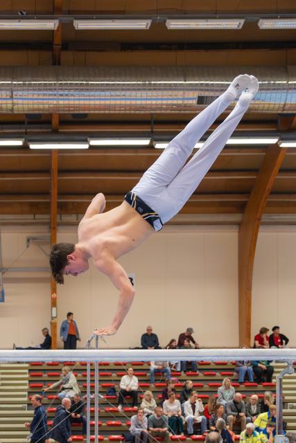 Young male gymnast performs a straight horizontal hold on parallel bars, body perfectly rigid, in a packed indoor sports hall.