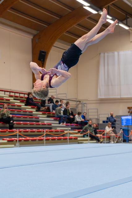 Young gymnast captured mid-backflip, fully inverted in the air, arms tucked, legs extended, in an indoor gymnastics hall.