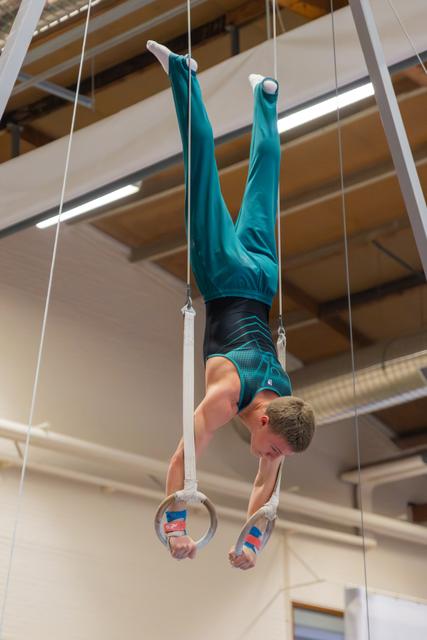Young male gymnast holds an inverted position on still rings, face taut with concentration, in a gymnasium.