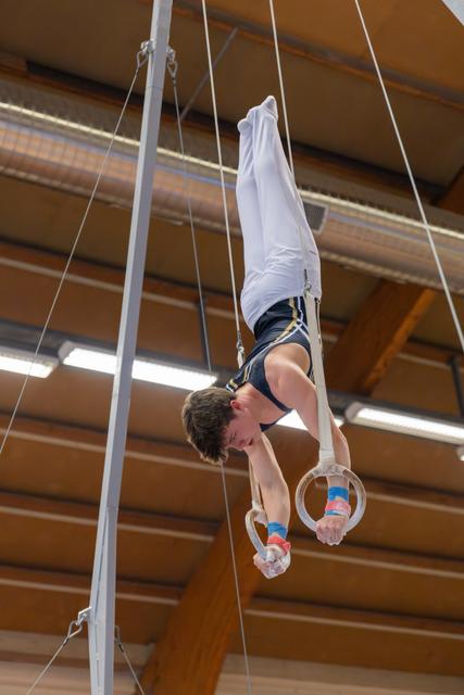 Young male gymnast performs an inverted hang on still rings, body fully extended upward in a gymnasium.