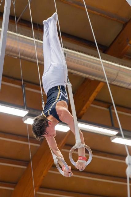 Young male gymnast performs an inverted hold on still rings, body perfectly straight, expression intense and focused, indoor gym.