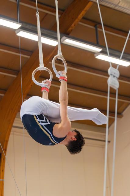 Young male gymnast hanging inverted on still rings, body curled, showing intense concentration in an indoor gym.