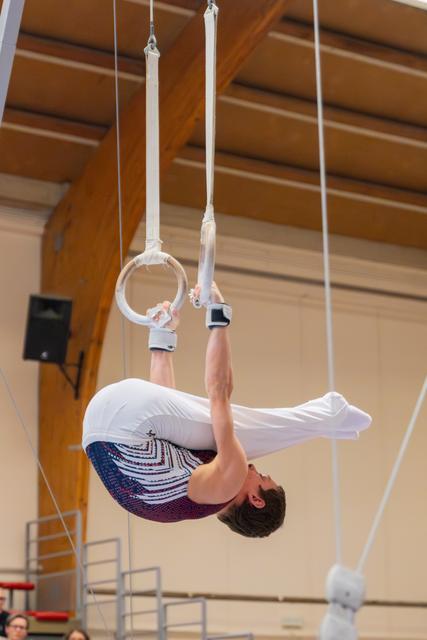 Male gymnast holds an inverted tuck position on still rings, arms straining with focused intensity in an indoor gymnasium.