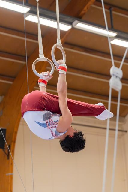 Young male gymnast hangs inverted on still rings, body arched with focus and control in an indoor gymnastics hall.
