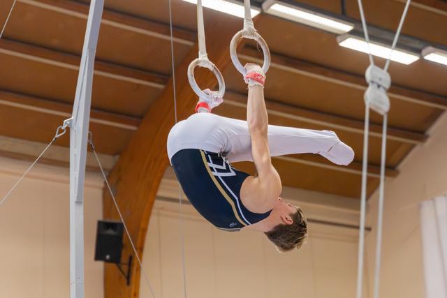 Young male gymnast performs an inverted tuck on still rings, wearing a navy leotard in an indoor gymnasium.