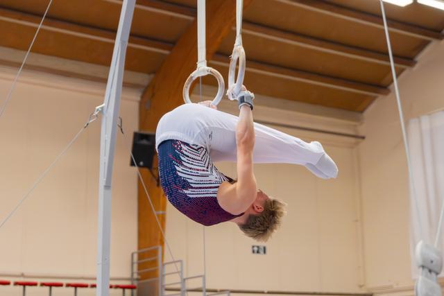 Young male gymnast performs an inverted tuck hold on still rings, displaying strength and control in an indoor gymnasium.