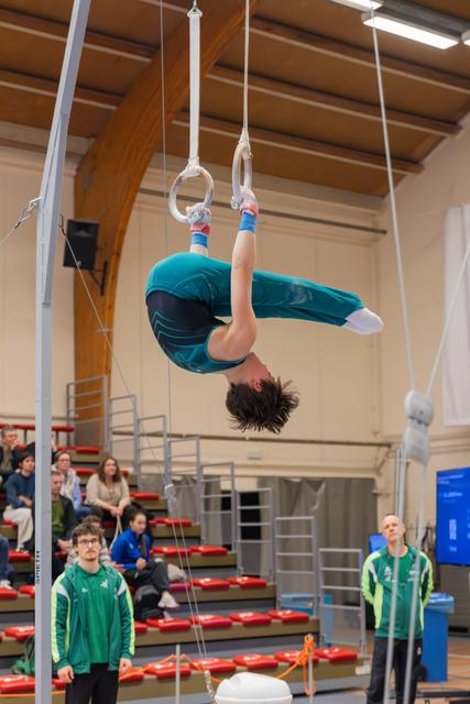 Young male gymnast hangs inverted on still rings, body arched, coaches watching below in indoor sports hall.