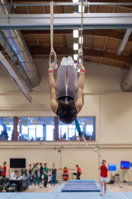 Gymnast hangs inverted on still rings, legs together, showing control and focus in a bright indoor gymnasium.