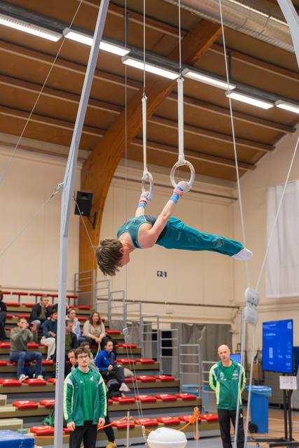 Young gymnast holds a dramatic iron cross position on still rings, body perfectly horizontal, in a packed indoor gymnasium.