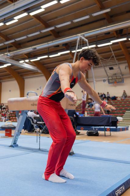 Young male gymnast landing on blue mat, leaning forward with arms extended, focused expression in indoor gymnastics hall.