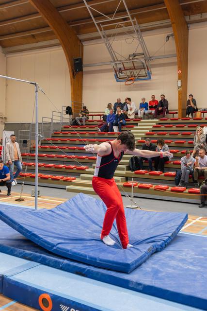 Young male gymnast lands on blue mat, arms wide for balance, leaning forward with intense focus. Spectators watch from bleachers.