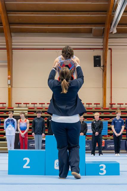 Official places a medal around a gymnast's neck on the first-place podium, as other competitors watch in a gymnasium.