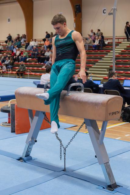 Young male gymnast steps onto a Spieth pommel horse, focused and composed, at an indoor gymnastics competition.
