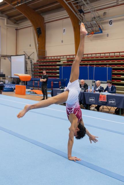 Young gymnast executes a one-armed handstand on the floor exercise mat during a gymnastics competition, legs split wide.
