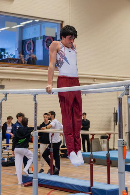 Young male gymnast grips parallel bars with focused concentration, wearing white leotard and red pants in a busy gym.