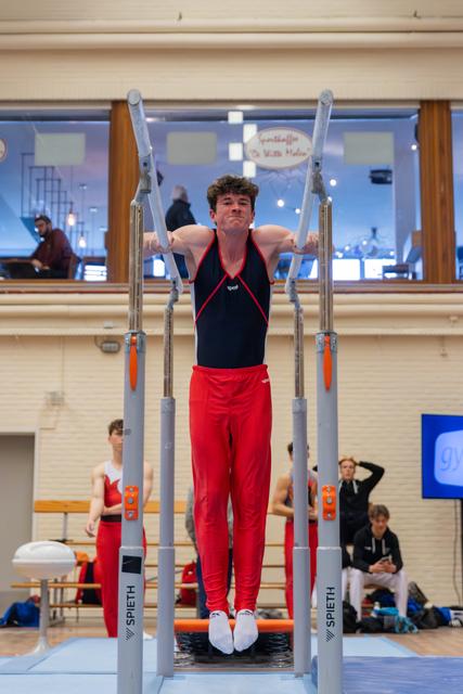 Young male gymnast grips parallel bars mid-routine, face showing intense concentration, wearing red and black leotard indoors.