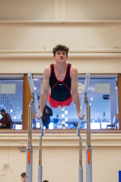 Young male gymnast holds a support position on parallel bars, focused expression, indoor gymnastics hall.