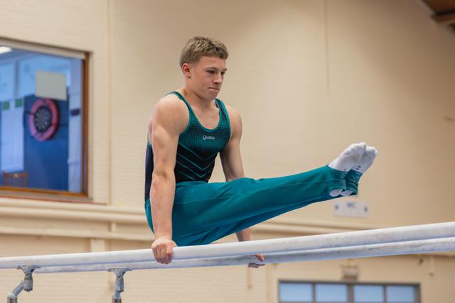 Young male gymnast performs a straddle hold on parallel bars, focused expression, teal leotard, indoor gym.
