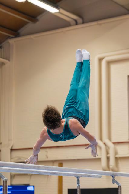 Young male gymnast performs a handstand on parallel bars, legs fully extended upward, wearing teal leotard and pants.