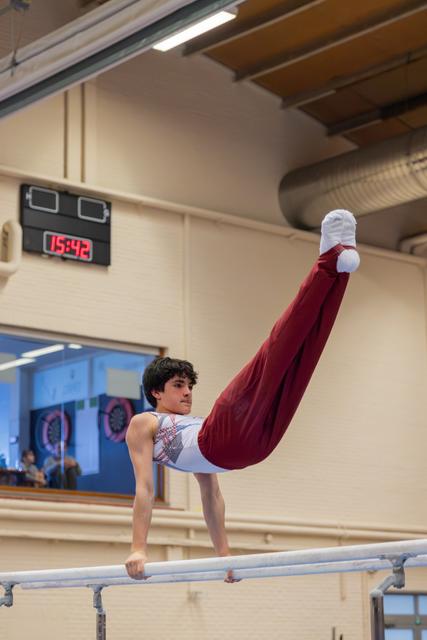 Young male gymnast performs a dramatic L-sit hold on parallel bars, legs raised high, intense focus in a competition gym.