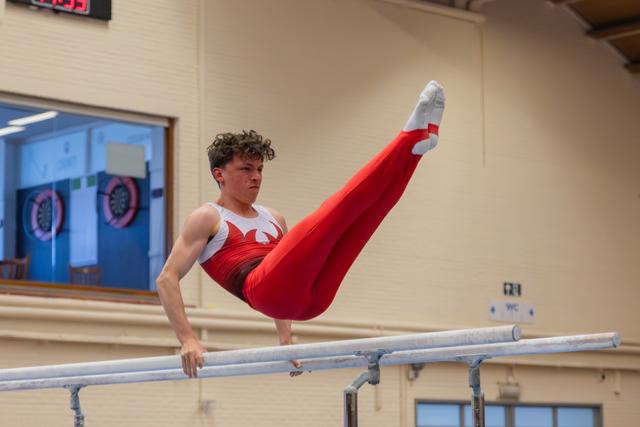 Young male gymnast holds a straddle L-sit on parallel bars, legs raised high, expression intense and focused.
