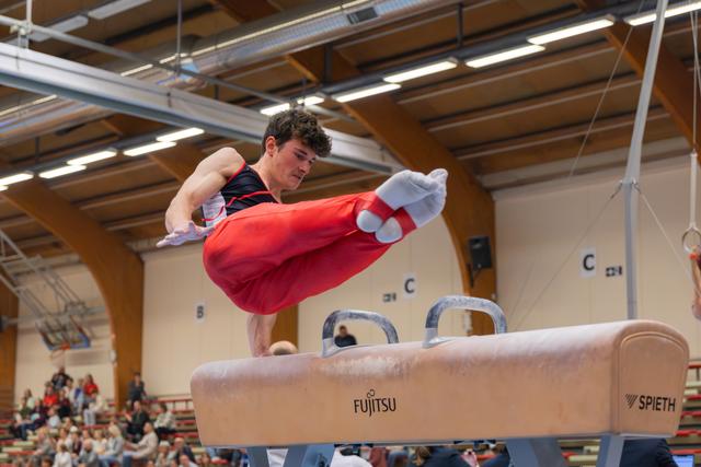 Young male gymnast swings his legs powerfully over a Spieth pommel horse, focused expression, indoor gymnastics hall.
