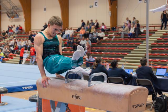 Young male gymnast performs on a Fujitsu-branded pommel horse, legs raised, concentrating intently during a gymnastics meet.