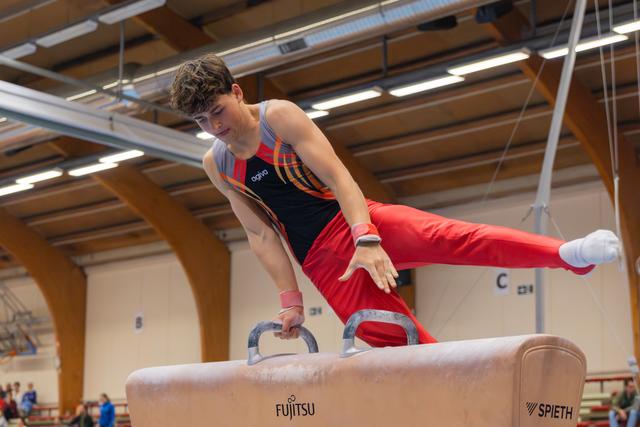 Young male gymnast performs on pommel horse, leaning forward with intense focus, legs extended mid-routine in indoor gym.