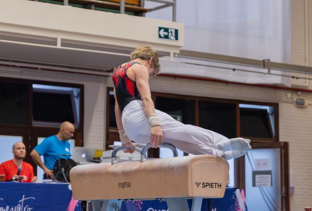 Young male gymnast performs pommel horse routine, leaning forward with intense focus, at an indoor competition venue.