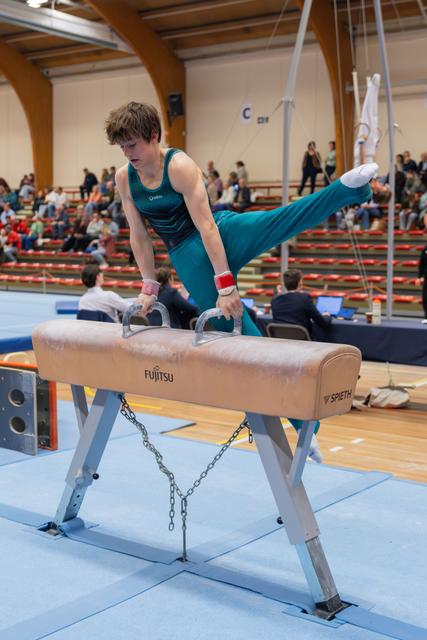 Young male gymnast in teal leotard performs leg extension on pommel horse, focused expression, indoor arena with spectators.