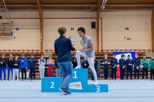 Young male gymnast on 1st place podium receives trophy from presenter, smiling warmly at a Gymfed awards ceremony.