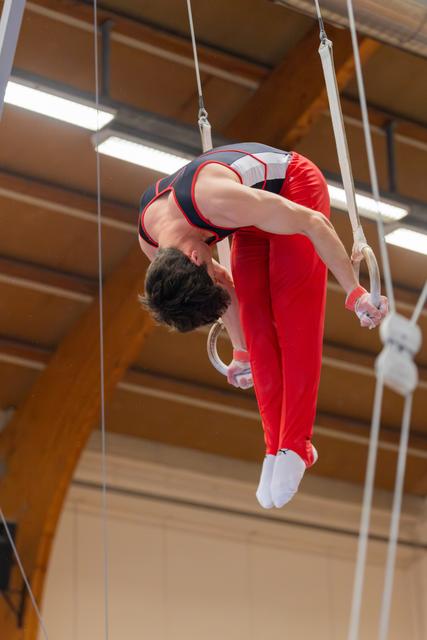 Male gymnast in red and black leotard performs a dramatic backbend on still rings in an indoor gymnasium.