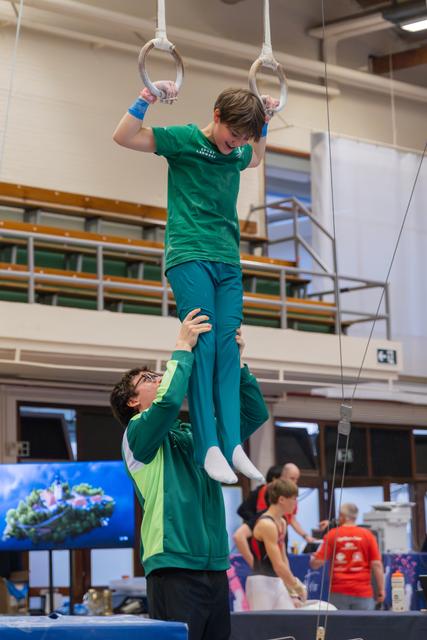 Smiling boy in green hangs from gymnastics rings while a coach steadies his legs in a busy sports hall.