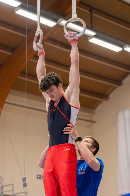 Young male gymnast grips still rings with intense focus while coach Simon supports his waist in an indoor gym.