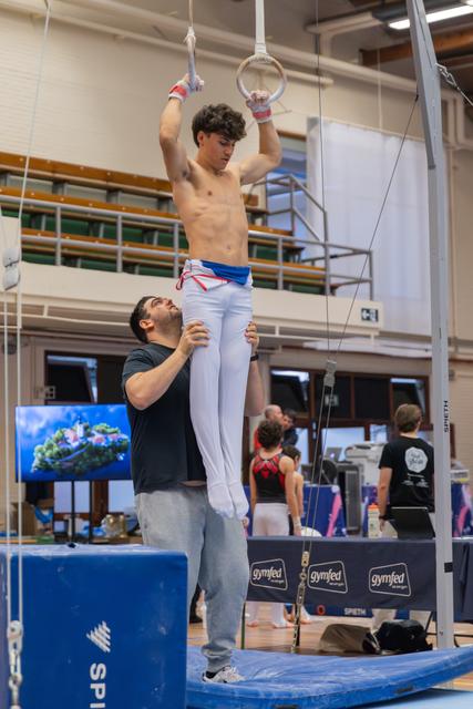 Shirtless male gymnast hangs focused on still rings while coach below steadies his legs in a training hall.