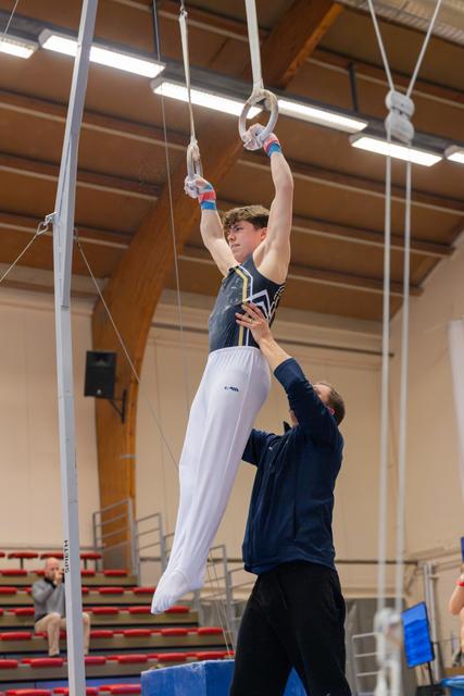 Young male gymnast holds still rings with focused expression while coach spots him below in an indoor gymnasium.