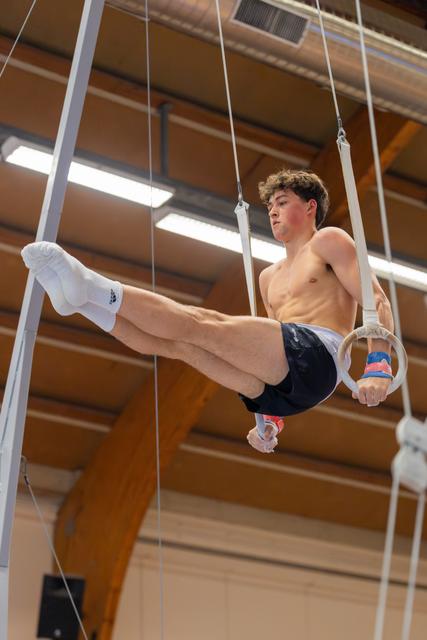 Young male gymnast performs a tucked L-sit on still rings, face set with intense concentration in an indoor gymnasium.