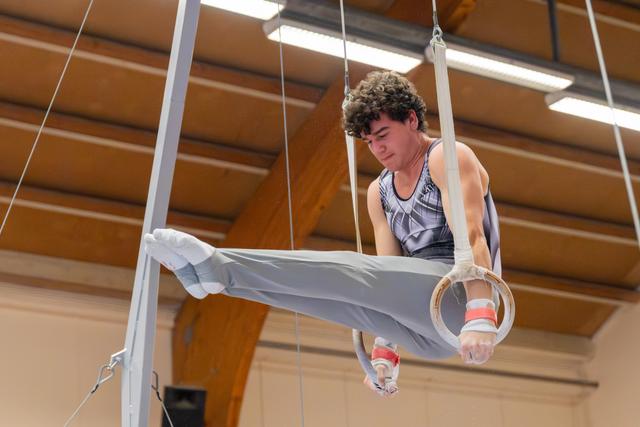 Young male gymnast with curly hair performs a straddle hold on rings, focused expression, in a wooden-ceiling gymnasium.