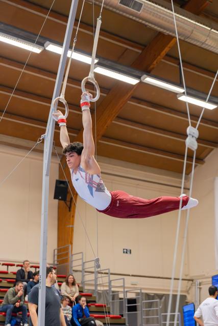Young male gymnast holds a pike position on still rings, intense focus, competing in an indoor sports hall.