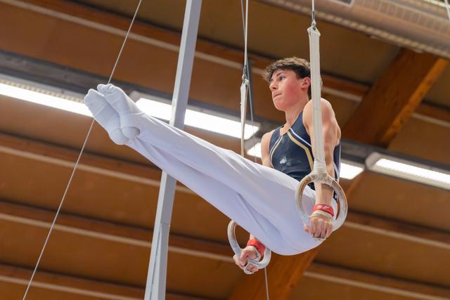 Young male gymnast performs an L-sit on still rings, legs raised, face focused and determined, indoor gymnasium.