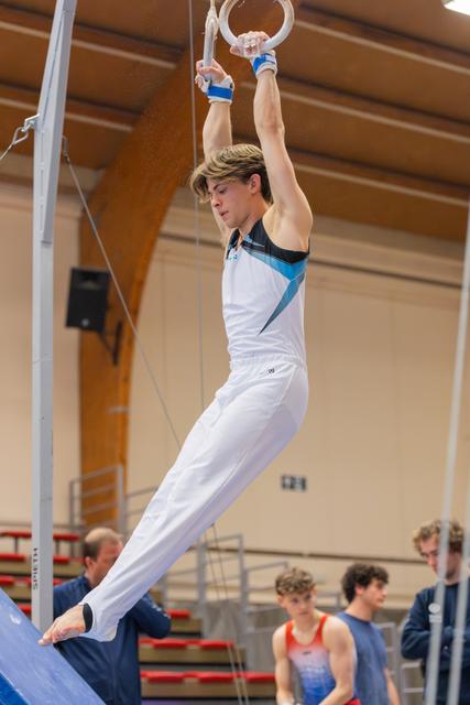 Young male gymnast hangs from still rings with focused expression, legs slightly raised, wearing white and blue leotard.