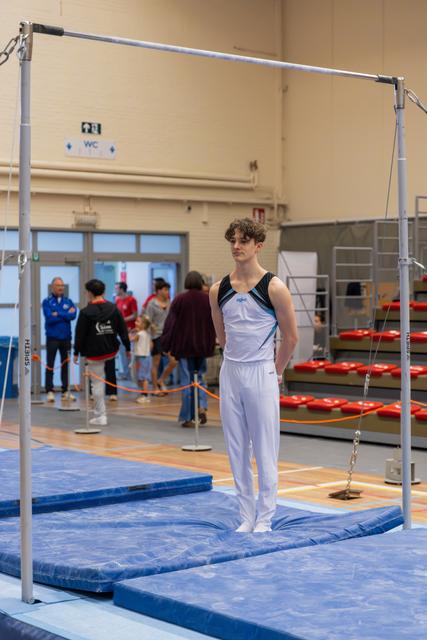 Young male gymnast stands composed on blue mat beneath the horizontal bar, gazing ahead before his routine.