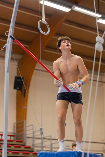 Young male gymnast stands on apparatus, gripping a red bar, looking upward with focused concentration in a gymnasium.